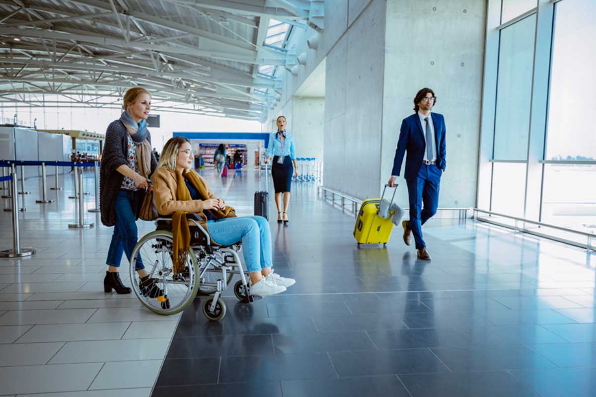 Navigating-Air-Travel-with-Rare-Disease-Disability A woman seated in a wheelchair, with another woman pushing the wheelchair behind her, at an airport.