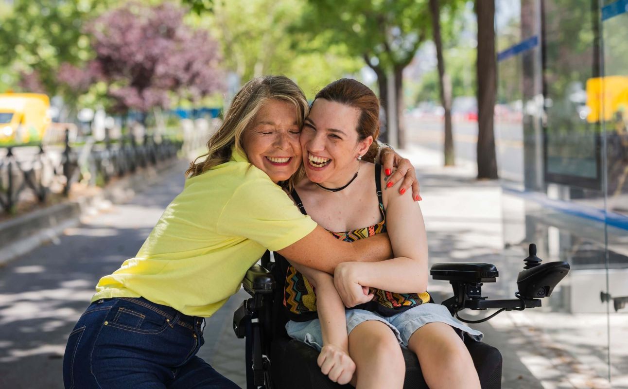 Two women smile and hug while sitting together outdoors on a sunny, tree-lined city street. One woman is seated in a wheelchair, and the other leans in affectionately beside her.